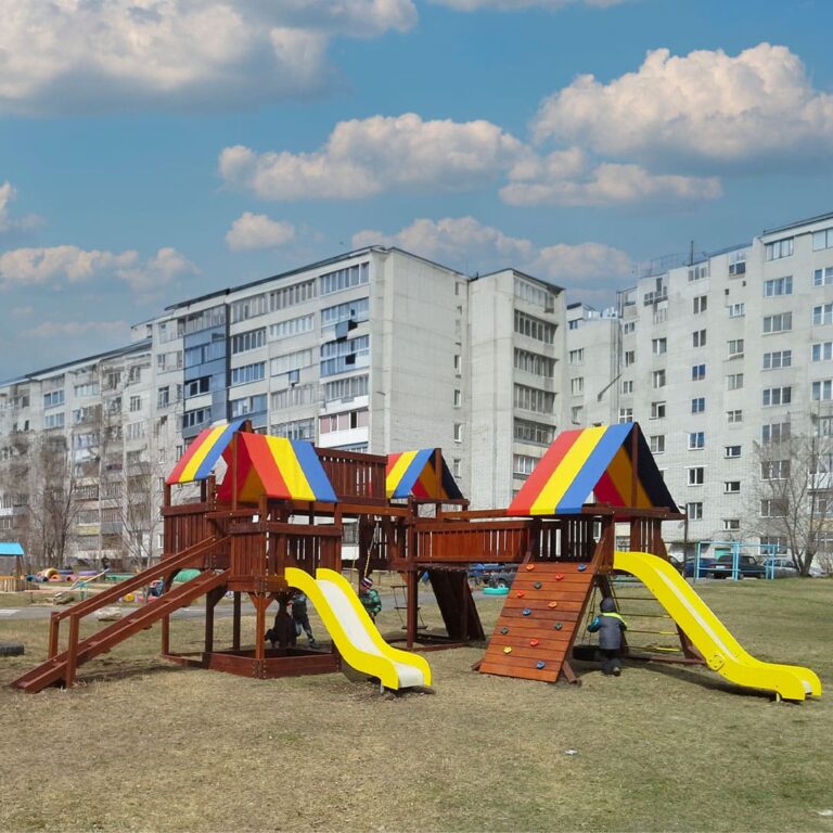 Небольшая детская игровая площадка из натурального дерева с качелями и горкой на фоне жилых многоквартирных домов